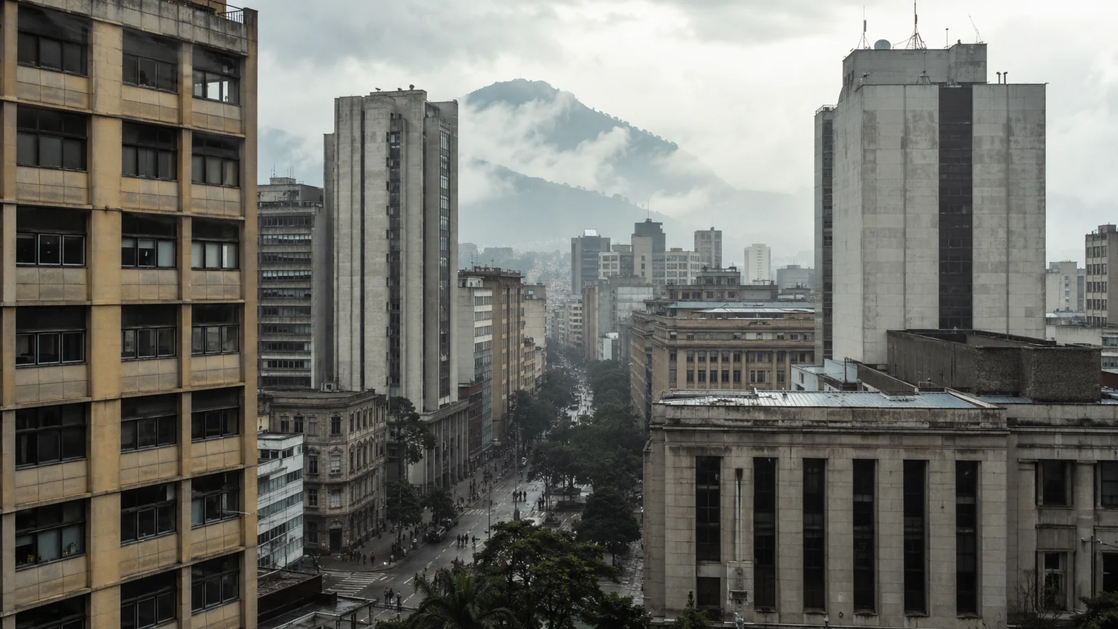 Colombian city centre under low cloud, mid-century concrete office towers with a forested mountain ridge behind